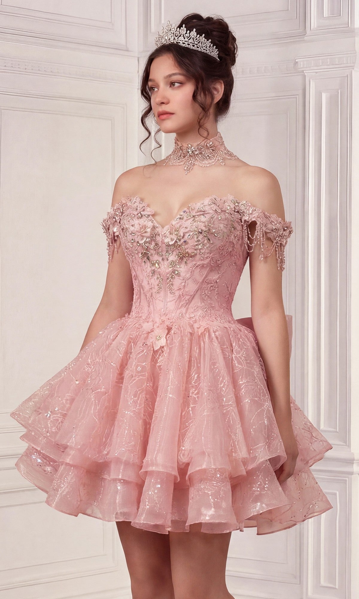 A woman in the Quinceanera Dress 15742 By Ladivine, featuring pink off-the-shoulder style, detachable skirt, floral embroidery, and a tiara, stands against an ornate white wall.